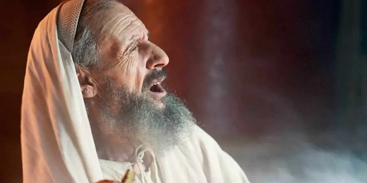 An elderly man, possibly Zechariah in the Bible, with a grey beard and wearing a white robe and head covering, cries out with his mouth open.