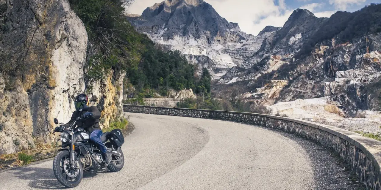 Un motociclista percorre una strada tortuosa tra le montagne della Toscana, con cave di marmo sullo sfondo.