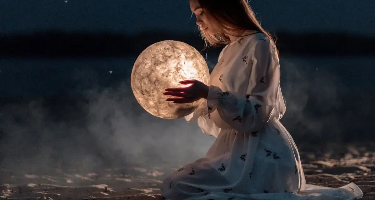 Dziewczyna w białej sukni trzyma w dłoniach świecącą kulę przypominającą księżyc. Nocne niebo, gwiazdy, mgła. Ascendent co to?