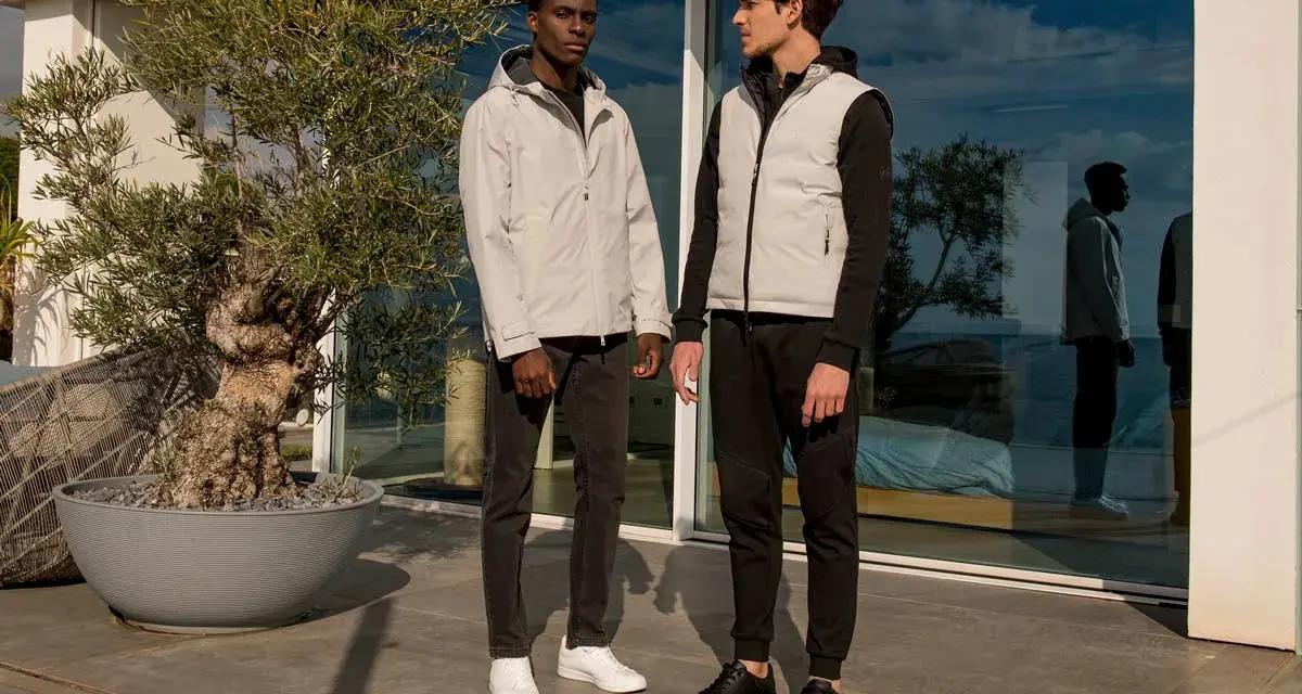Due uomini con un moderno stile outfit uomo, uno con giacca grigia e jeans, l'altro con gilet grigio e pantaloni sportivi neri.