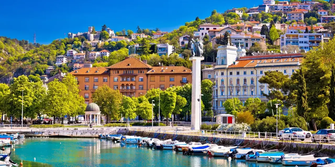 Malowniczy port w Chorwacji z łodziami przycumowanymi do nabrzeża. W tle widać wzgórze z zabudowaniami i zamek.