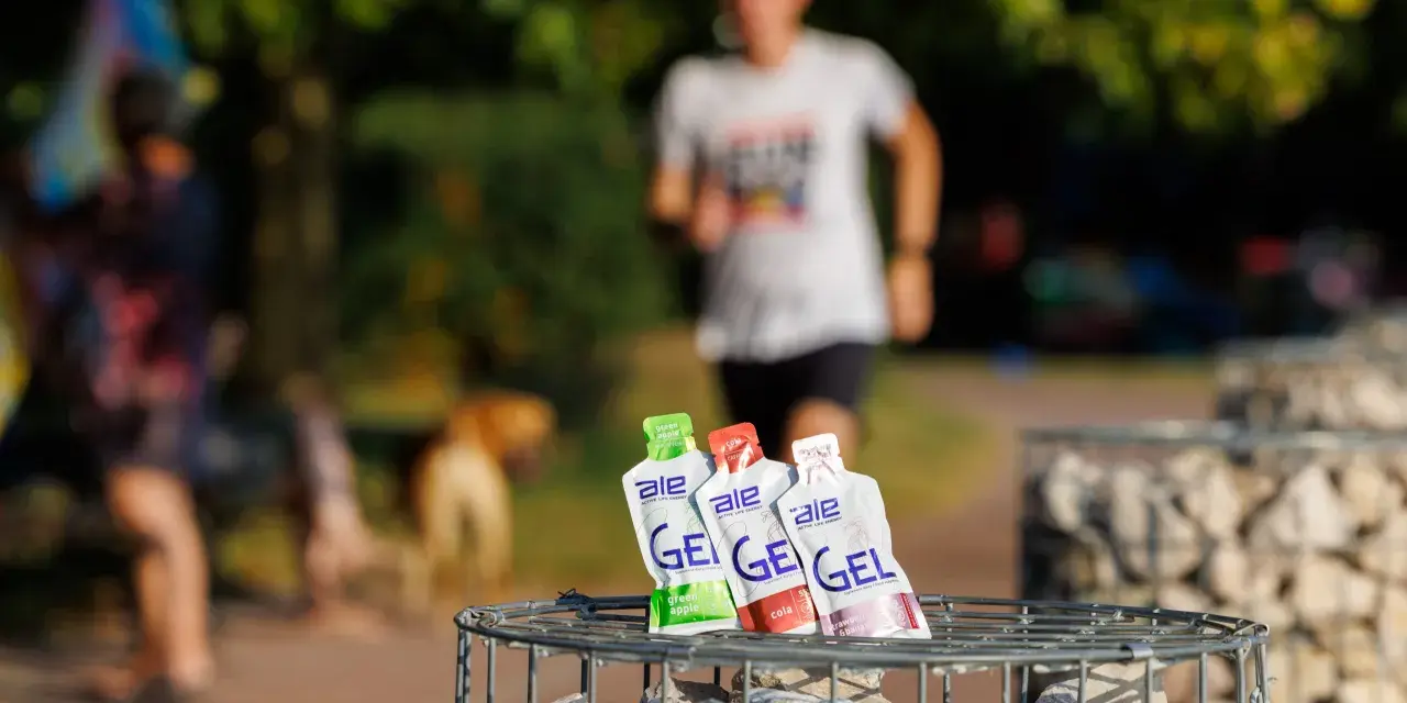 Trzy opakowania żeli energetycznych dla biegaczy: zielone jabłko, cola i truskawka z bananem, na tle biegnącej osoby.