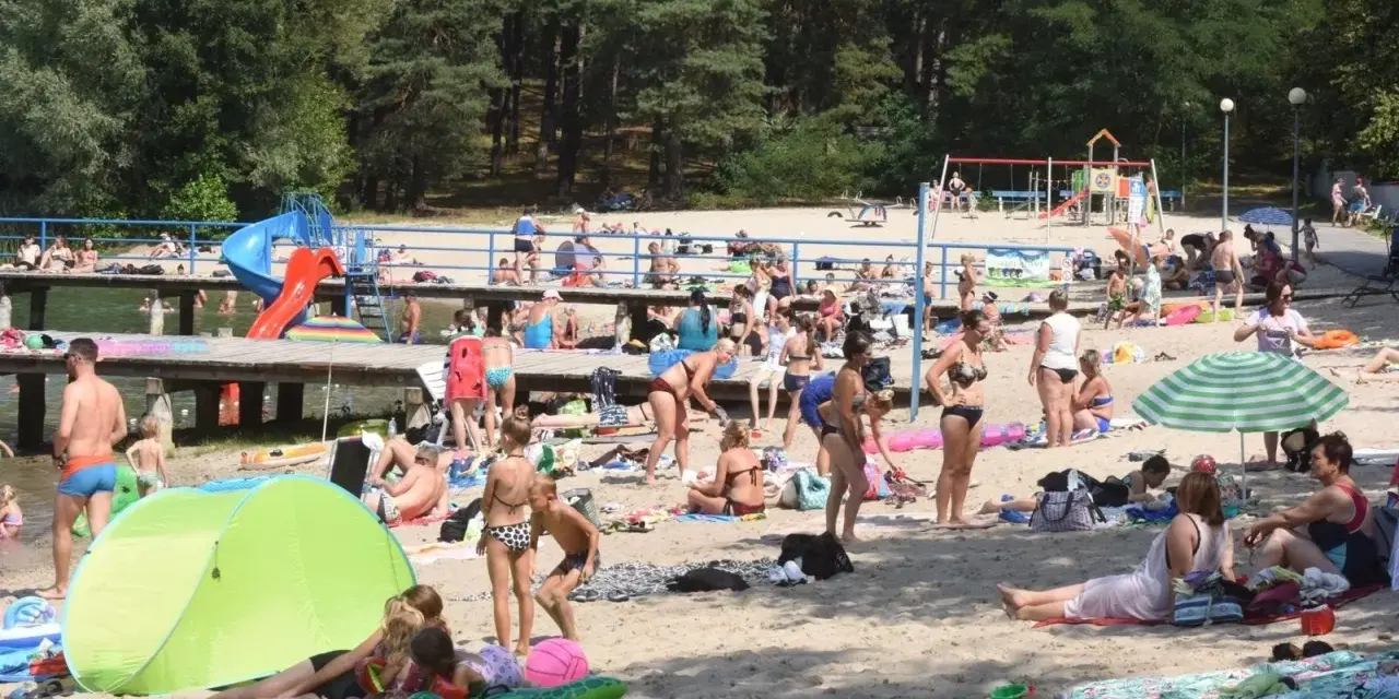 Tłoczna plaża nad jednym z **jezior blisko Wrocławia**. Ludzie opalają się, kąpią i odpoczywają na piasku.
