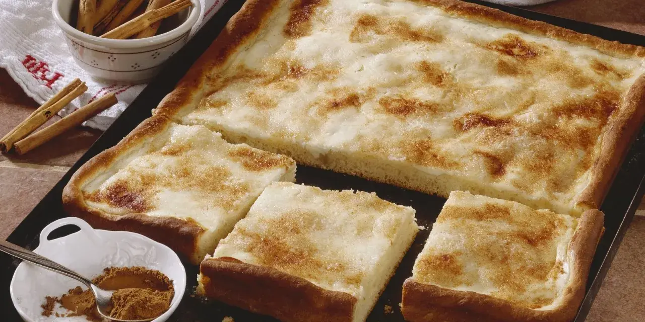 Ein köstlicher Rahmkuchen vom Blech, bestreut mit Zimt und Zucker. Daneben eine Schale mit Zimtstangen und Zimtpulver.