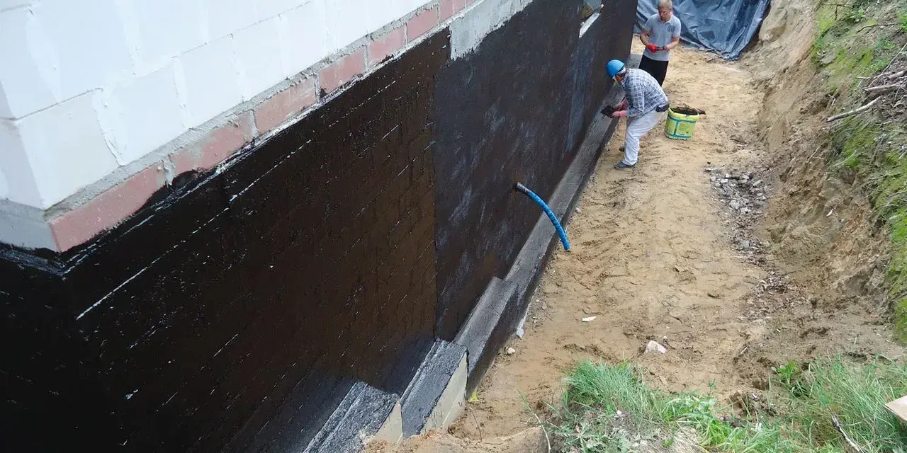Pracownicy nakładają masę KMB na fundamenty domu. Czarna powłoka chroni przed wilgocią.