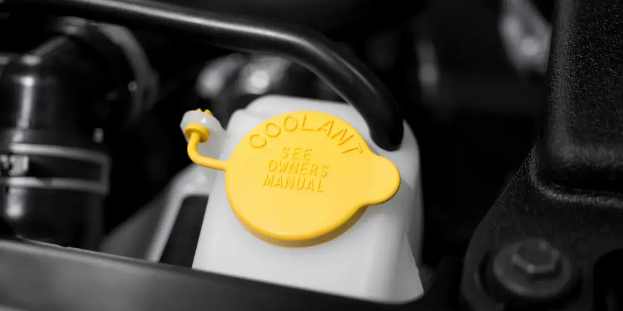 Close-up of a car's coolant reservoir with a yellow cap. The cap reads "COOLANT SEE OWNERS MANUAL," indicating where to find instructions on how to put coolant in car.