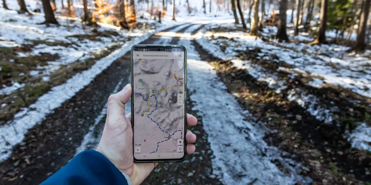 Telefon z mapą pokazuje szlak w lesie. Widać ślady śniegu na drodze, a na ekranie zaznaczony jest skopiec szlak.