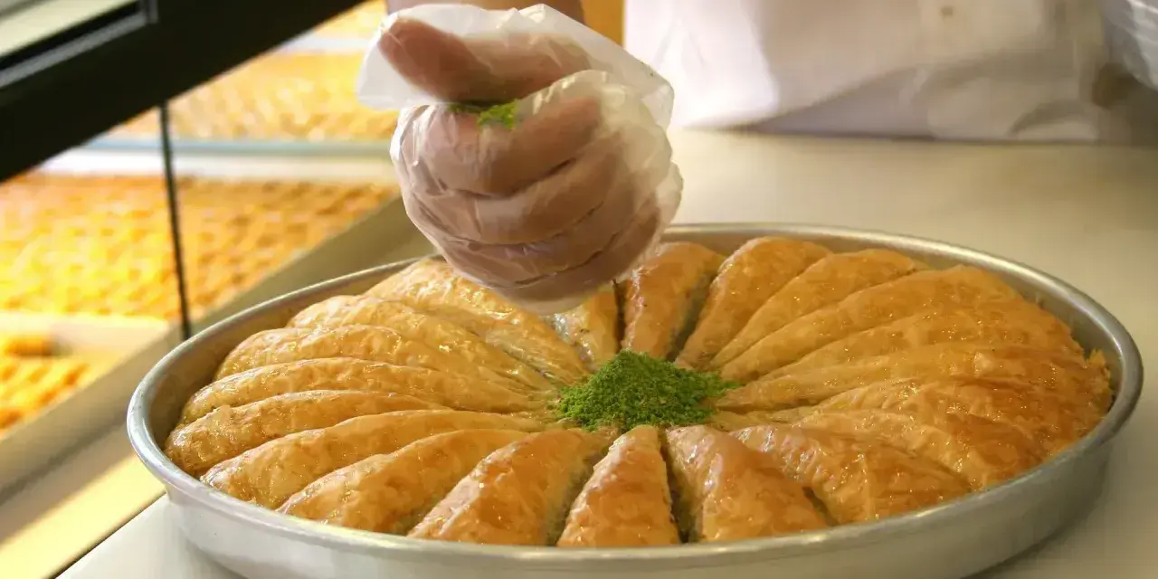 Ręka w rękawiczce posypuje zielonymi pistacjami okrągłą blachę z baklavą. To właśnie baklava co to za deser!