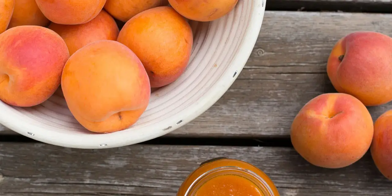 Ein Korb voller reifer Marillen, daneben ein Glas Marillenmarmelade. Was sind Marillen? Süße Früchte, perfekt für Marmelade!