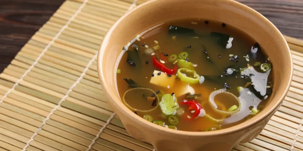 Aksamitna zupa miso z tofu, wodorostami, szczypiorkiem i papryczką chili, podana w ceramicznej miseczce na bambusowej macie.