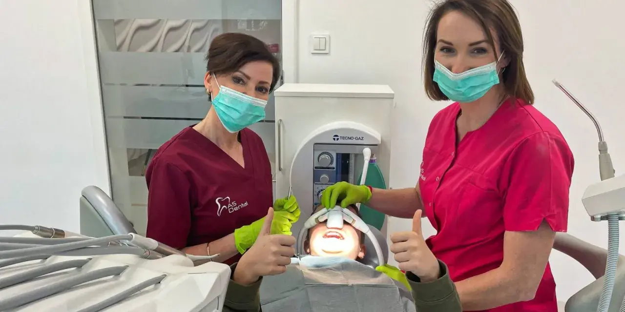 Dwie dentystki w maseczkach i rękawiczkach z uśmiechniętym pacjentem w gabinecie stomatologicznym.