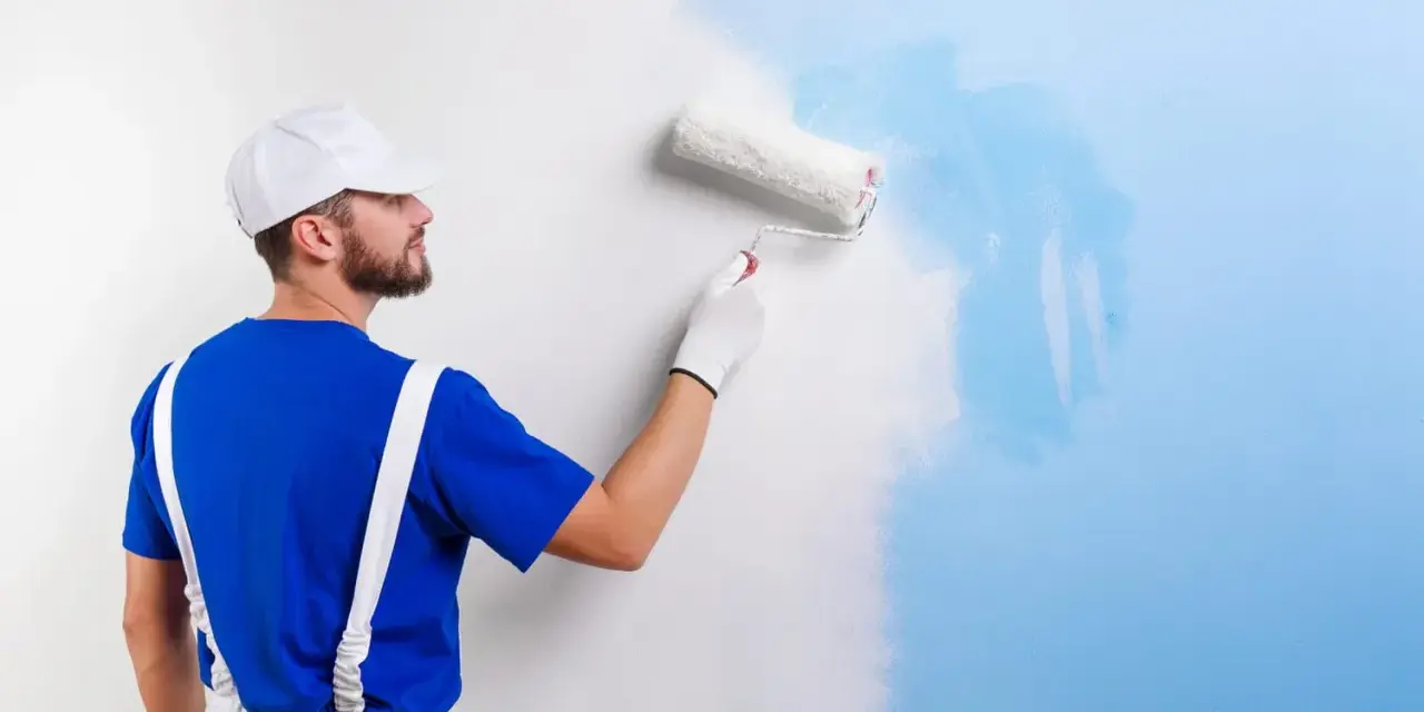 Un peintre en bleu et blanc applique une nouvelle couche de peinture bleue sur un mur blanc. C'est une peinture sur peinture en cours.