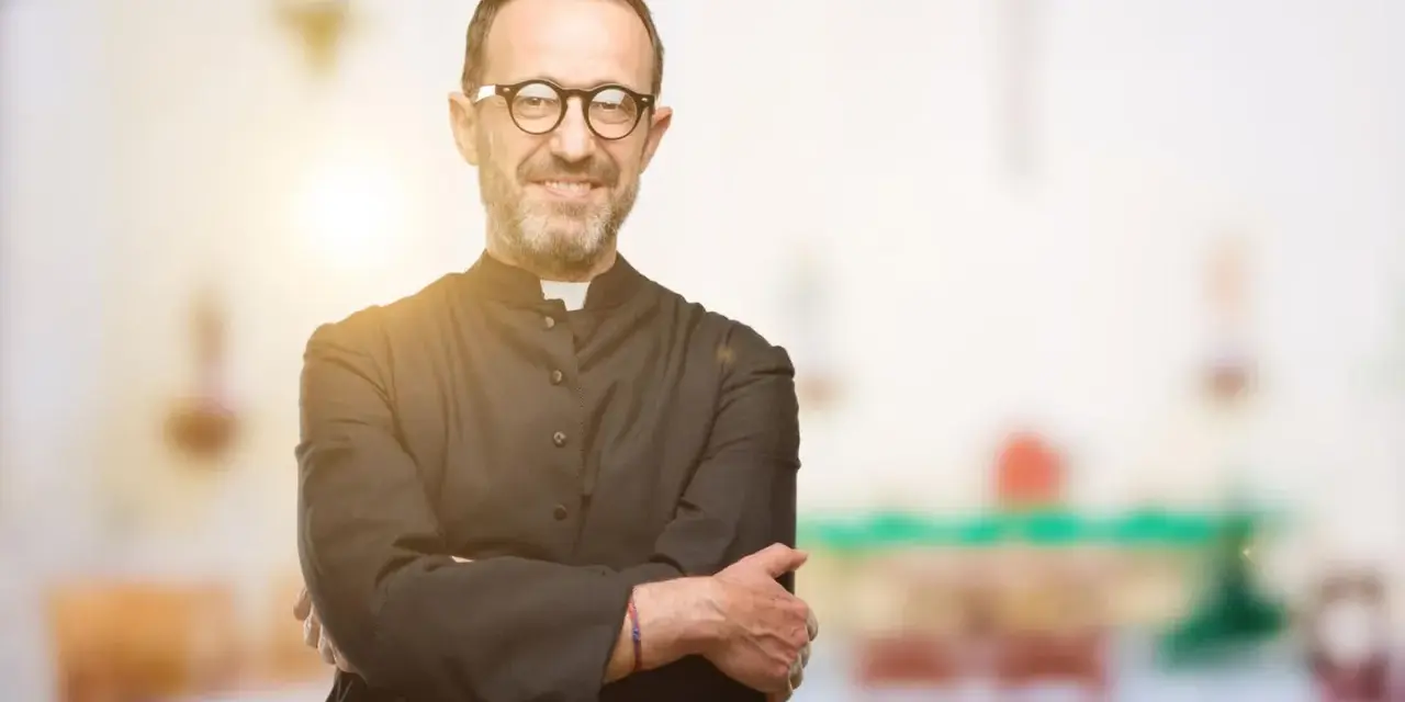 Ein lächelnder evangelischer Pfarrer mit Brille und überkreuzten Armen steht vor einem unscharfen Kircheninterieur.