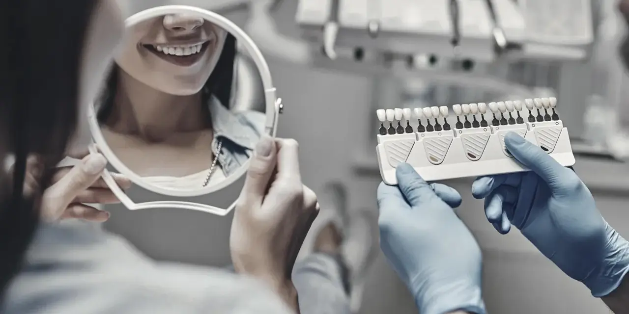 Dentysta pokazuje pacjentce wzornik kolorów zębów, pacjentka ogląda swój uśmiech w lusterku.