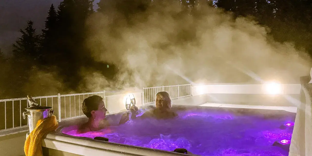 Zwei Frauen stoßen mit Champagner in einem beleuchteten Whirlpool an, umgeben von Dampf und Bäumen. Ein perfektes Wochenende mit Gott.