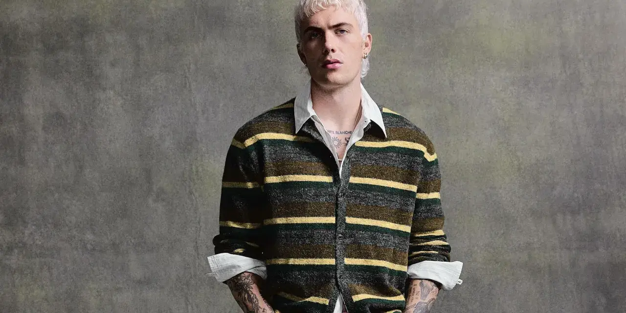 Joven con pelo rubio platino y tatuajes lleva un jersey de rayas verdes y amarillas, mostrando que **que jerseys se llevan este año** son los de estilo retro.