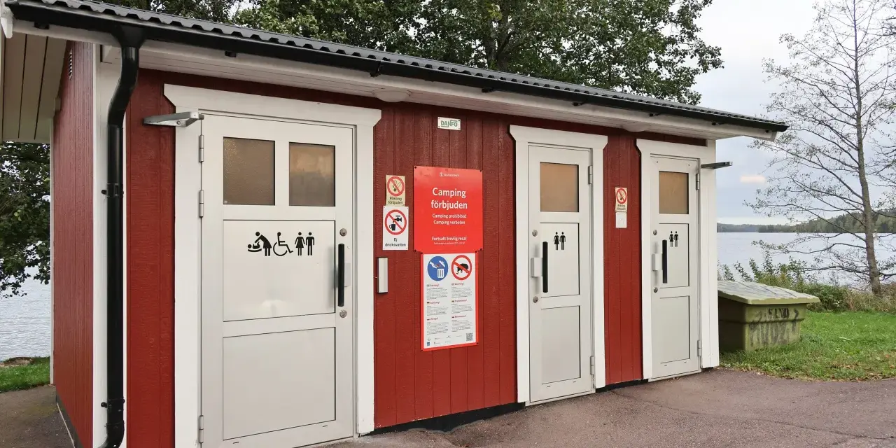 Röd byggnad med toaletter vid en sjö. Skyltar visar att camping är förbjuden. En trevlig rastplats med toalett för resenärer.