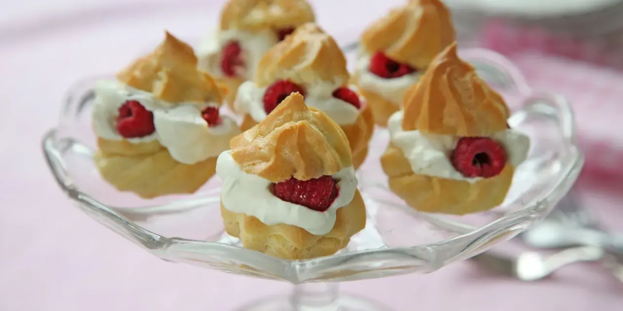 Små petit choux fyllda med grädde och hallon på ett glasfat.