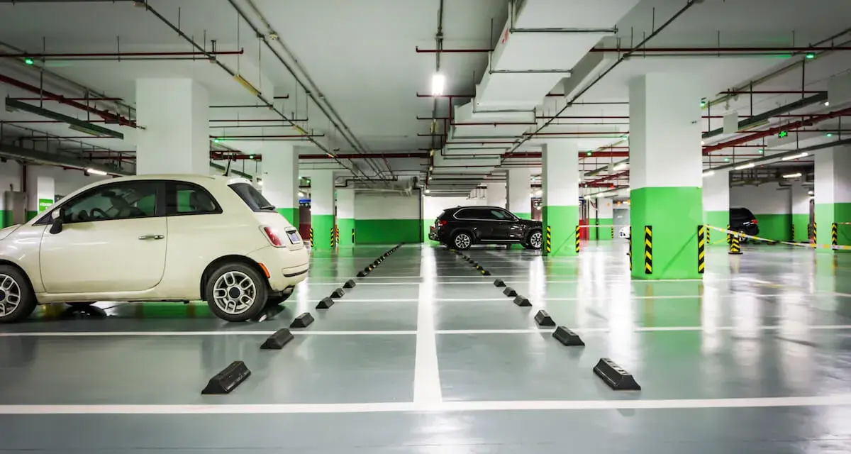 Jasny samochód i ciemny SUV stoją na parkingu podziemnym. Widać wyraźnie wyznaczoną szerokość miejsca parkingowego.