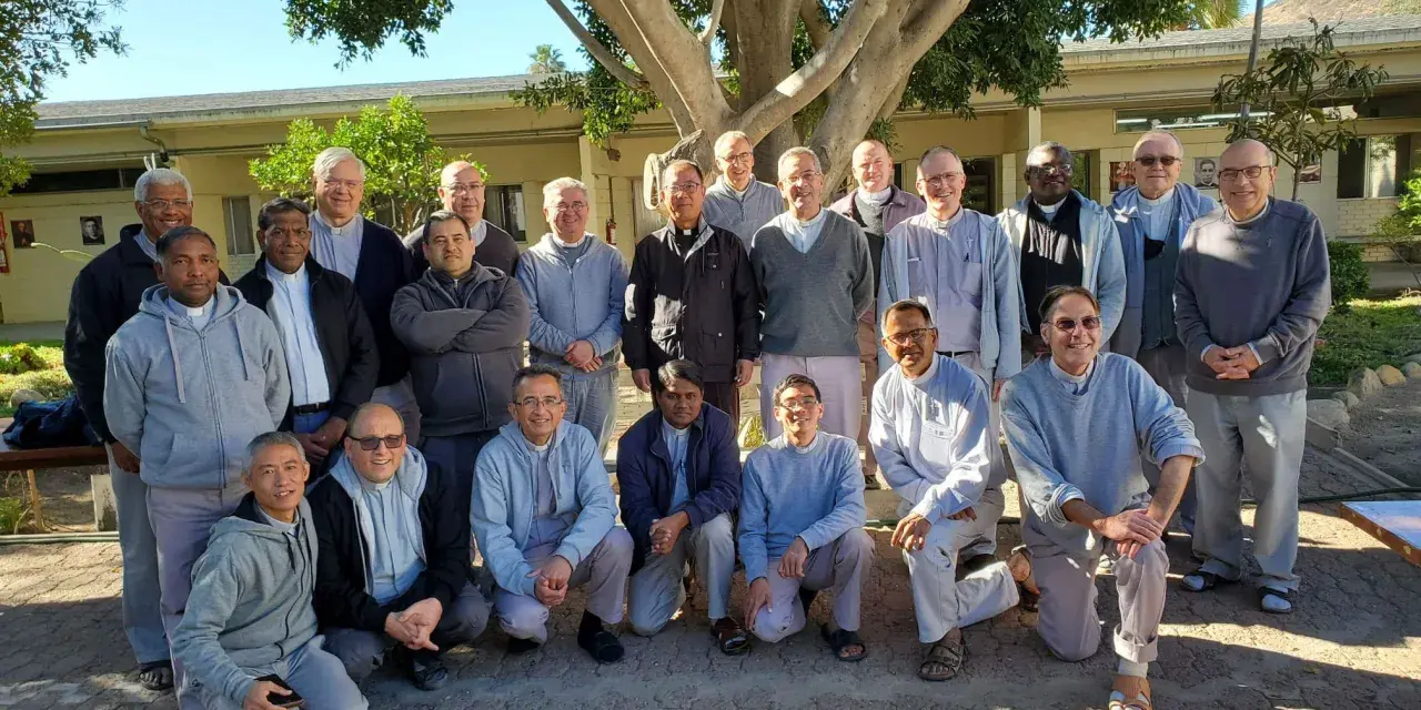 A group of missionaries of charity fathers pose for a photo outdoors, standing and kneeling under a large tree.