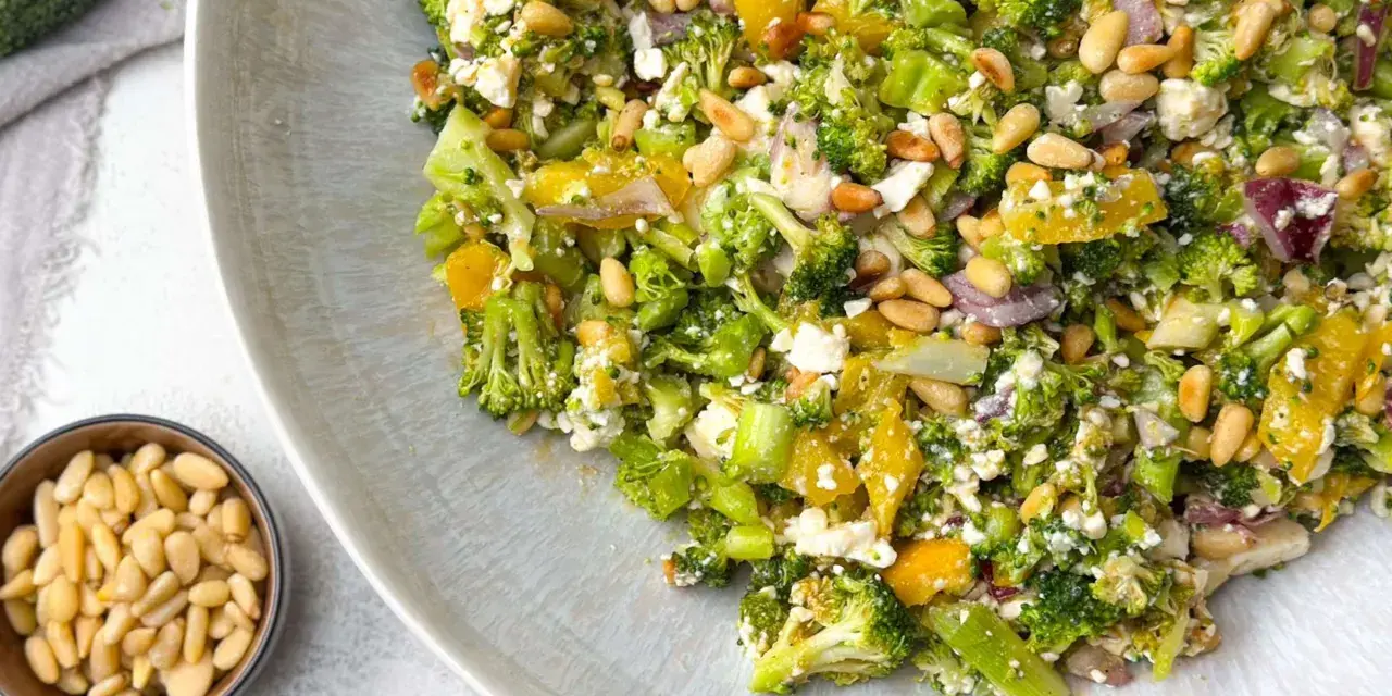 Ein bunter gekochter Brokkoli-Salat mit Pinienkernen, Feta und Orangenstücken auf einem Teller.