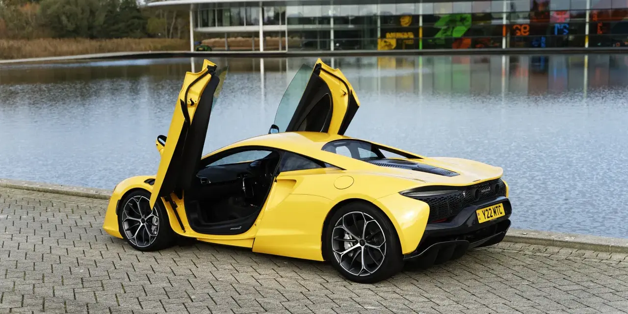 Żółty McLaren Artura z otwartymi drzwiami, stojący nad jeziorem przy nowoczesnym budynku.