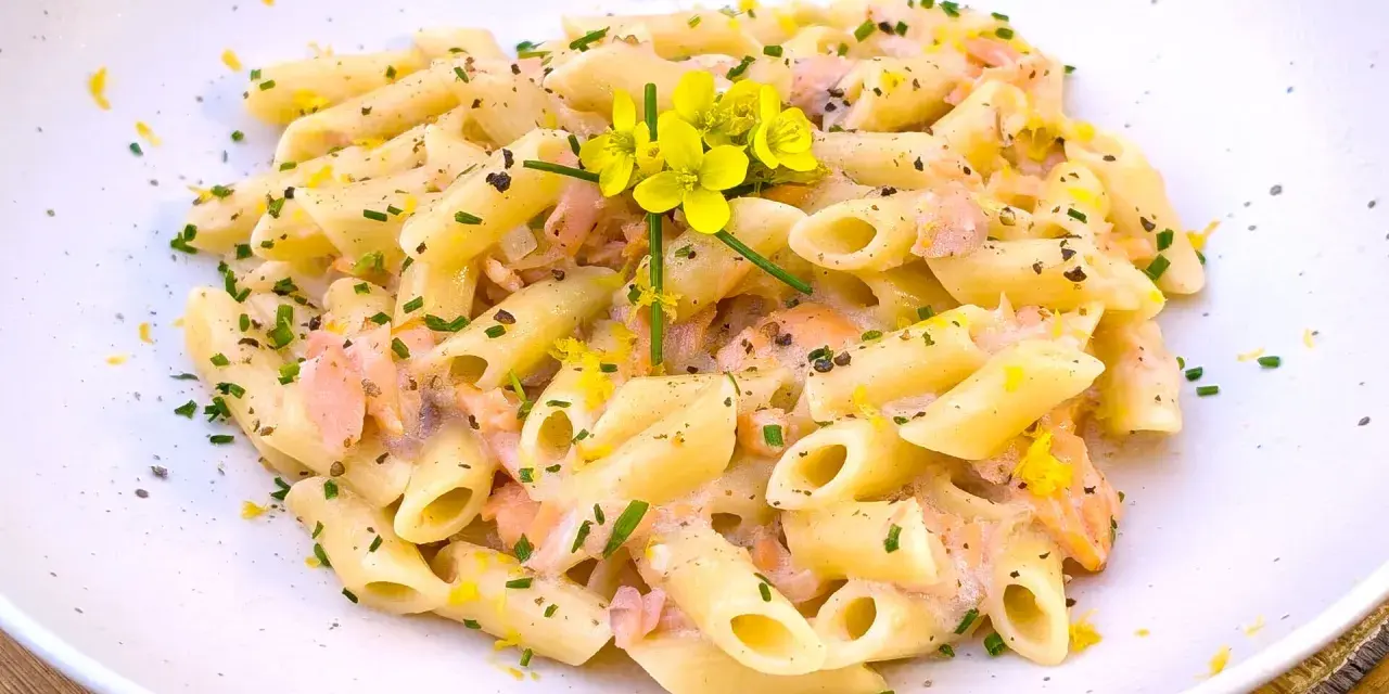 Pasta penne cremosa con trozos de salmón rosado, ralladura de limón y flores amarillas.
