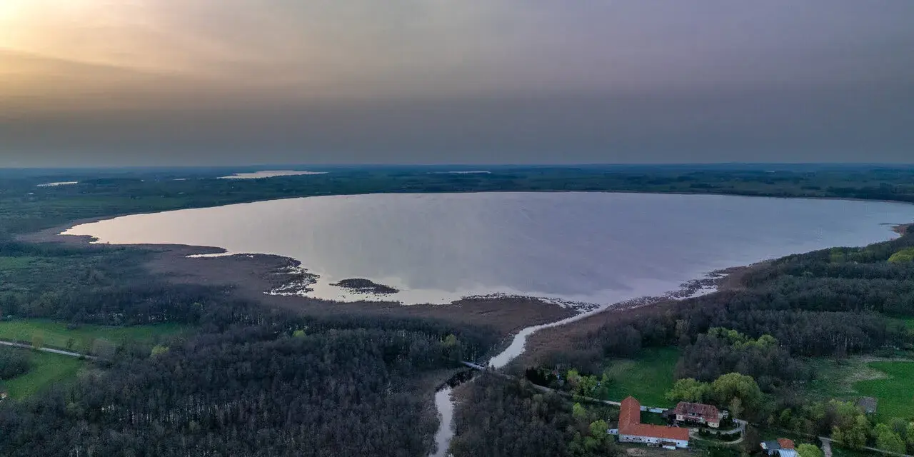 Rozległe jezioro Tuchlin otoczone lasem, z widocznym budynkiem i drogą. Zachód słońca maluje niebo.