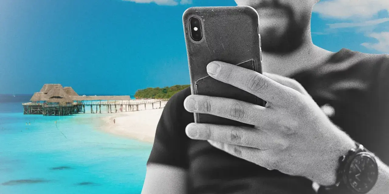 Mężczyzna z telefonem na plaży na Zanzibarze, sprawdzający zanzibar internet. W tle turkusowa woda i drewniany pomost.