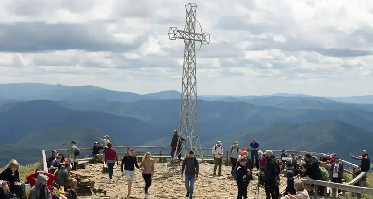 Ludzie odpoczywają na szczycie Tarnicy, podziwiając widok na góry. Metalowy krzyż góruje nad nimi.