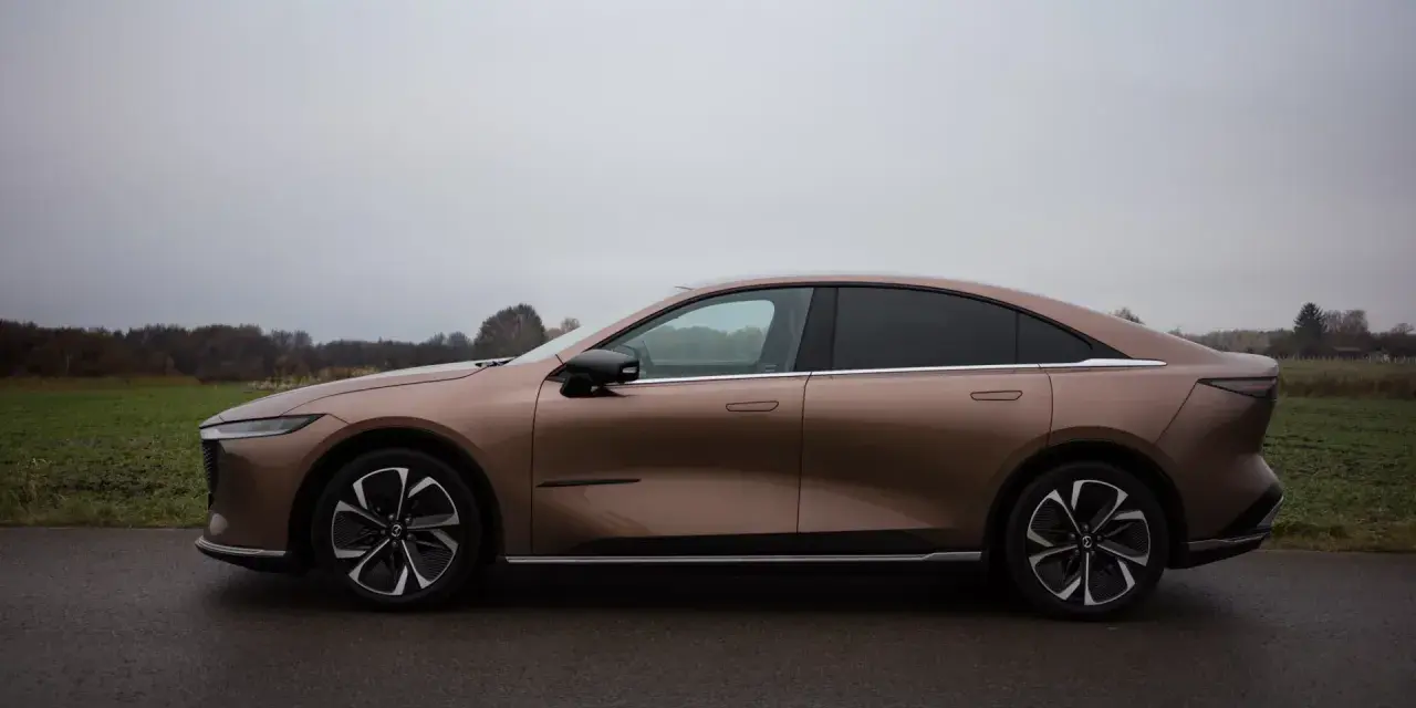 Brązowy samochód elektryczny Mazda6e widziany z boku na tle pochmurnego nieba i krajobrazu.