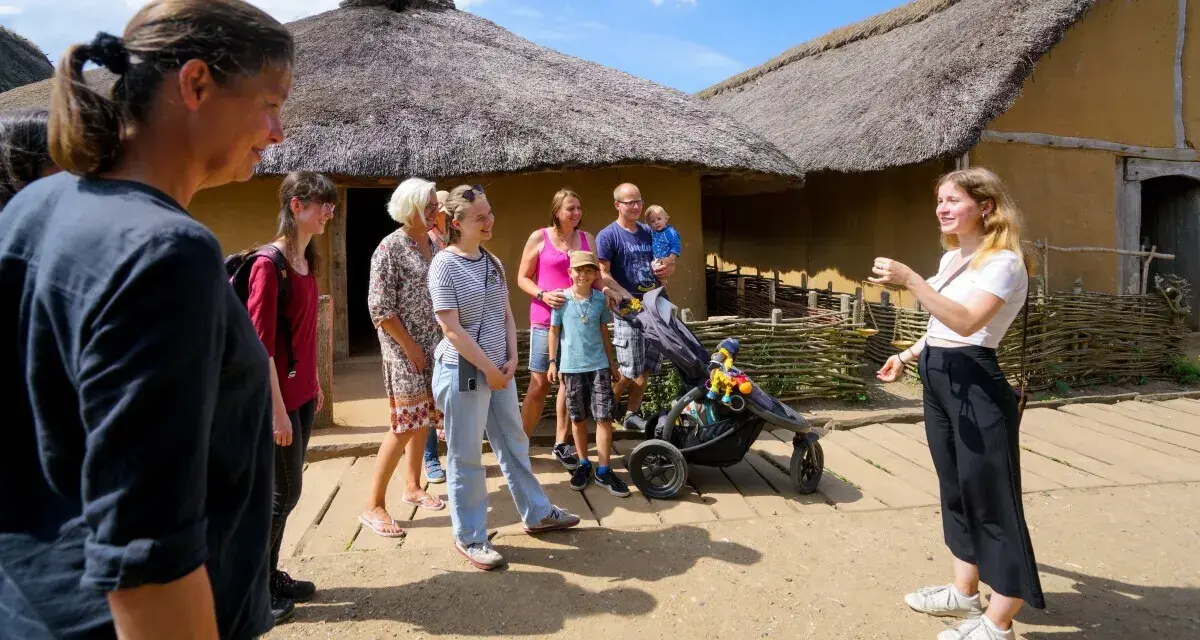 Familienurlaub in Haithabu: Eine Gruppe erkundet historische Häuser mit Strohdächern, während eine junge Frau eine Führung gibt.
