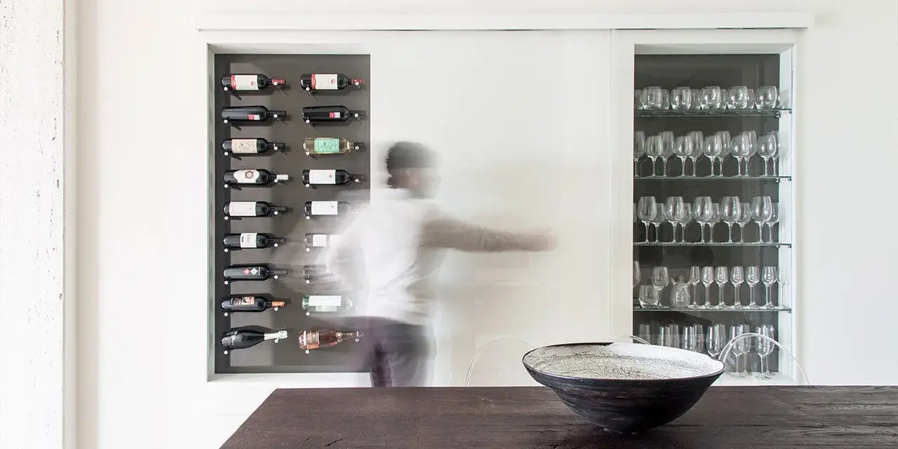 Una nicchia nel muro portante ospita una cantinetta per vini e una vetrina di bicchieri. Una figura sfocata si muove tra i due spazi.