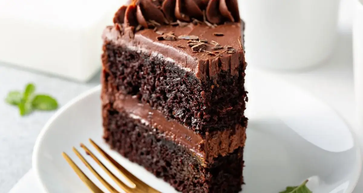 Rebanada de tarta de chocolate con glaseado cremoso y virutas de chocolate. Un tenedor dorado espera pacientemente para probar este delicioso postre.