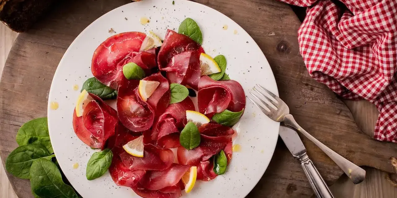 Bresaola affettata finemente, guarnita con spinacini freschi e spicchi di limone. Un'idea semplice su come condire la bresaola per un antipasto leggero.
