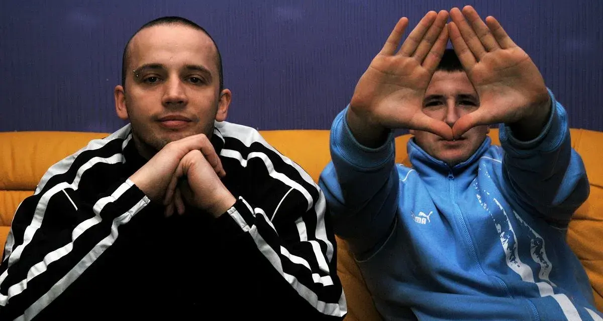 Dwóch mężczyzn w dresach, jeden z rękami złożonymi na brodzie, drugi tworzy rękami kształt serca. Klimat hip hop i rap.