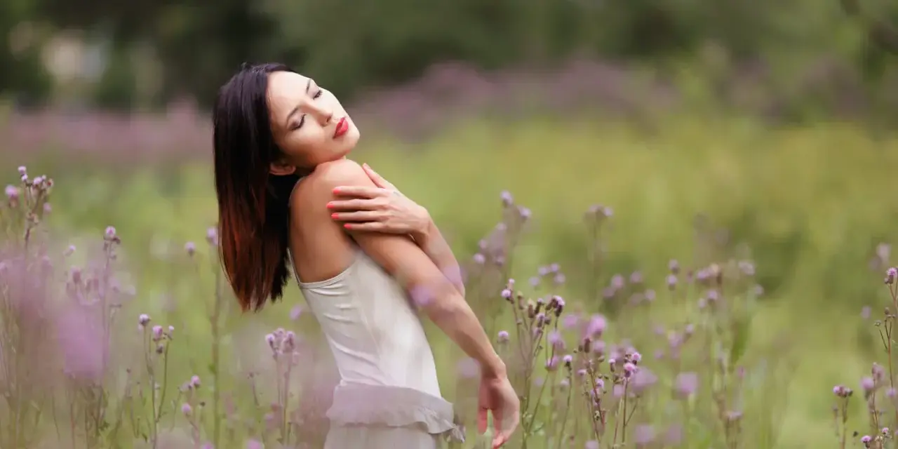 Portrait en bokeh d'une femme aux yeux fermés, bras croisés sur la poitrine, dans un champ de fleurs sauvages mauves.