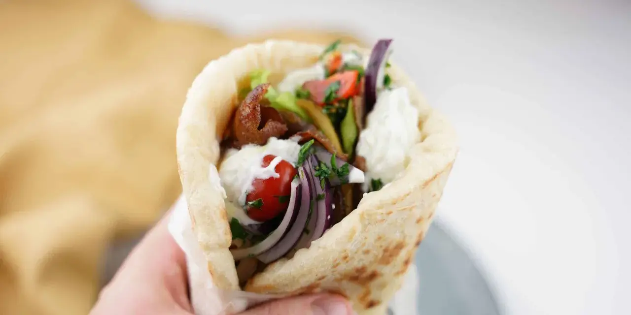 En hand håller en gyros med kött, grönsaker och vit sås. Perfekt för supergod fredagsmat!