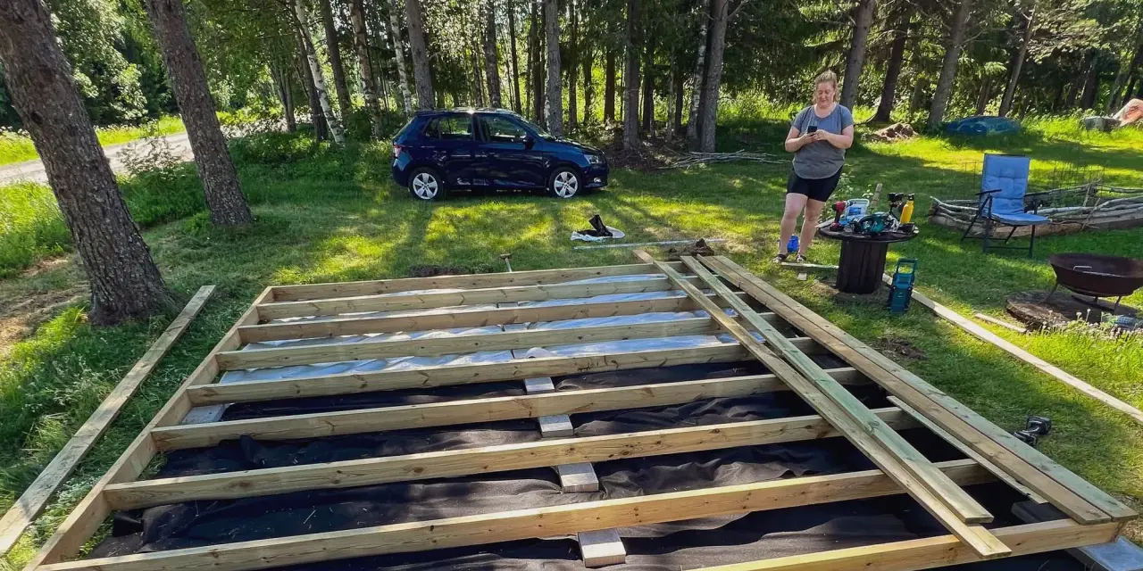 En kvinna förbereder sig för att skruva trall på en nybyggd altan i en lummig trädgård.