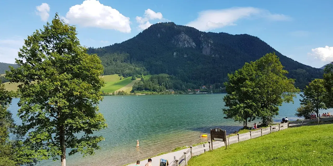 Ein sonniger Tag am bayrischzell see. Menschen spazieren am Ufer entlang, während im Hintergrund ein bewaldeter Berg aufragt.