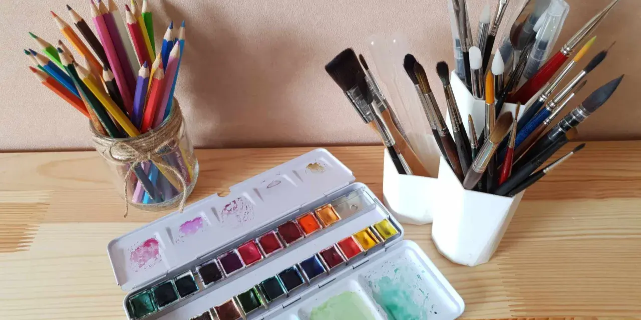 Une palette d'aquarelle ouverte, des crayons de couleur dans un bocal et plusieurs pinceaux indispensables aquarelle dans des pots blancs.