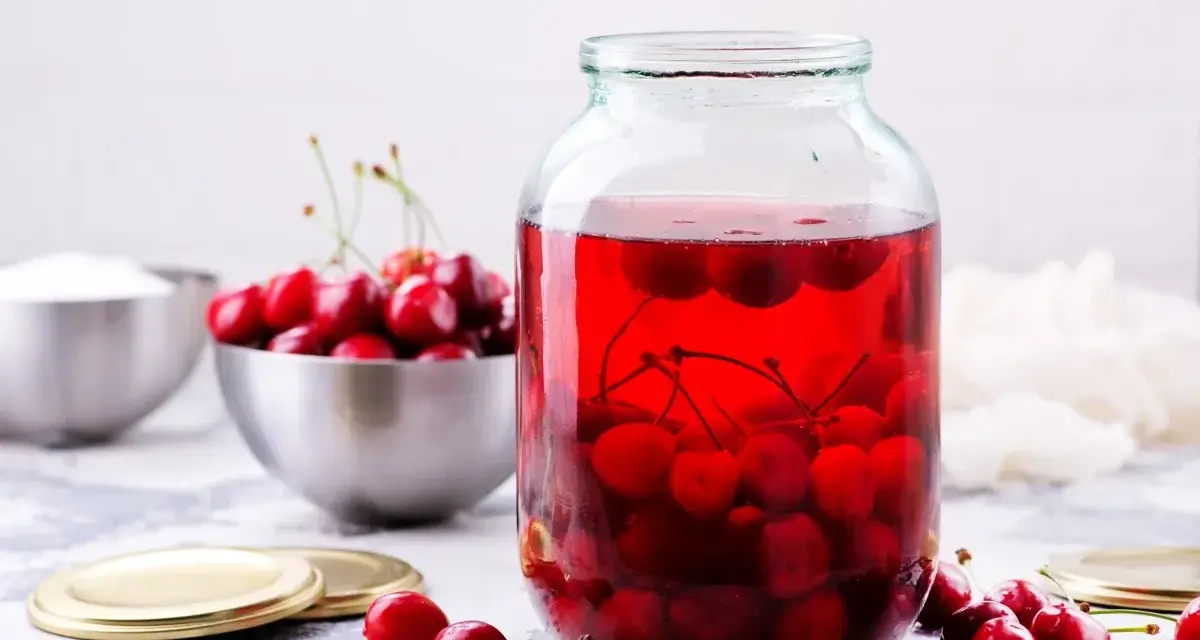 Duży słoik wypełniony rubinowym kompotem z wiśni, obok miska świeżych owoców i cukru.