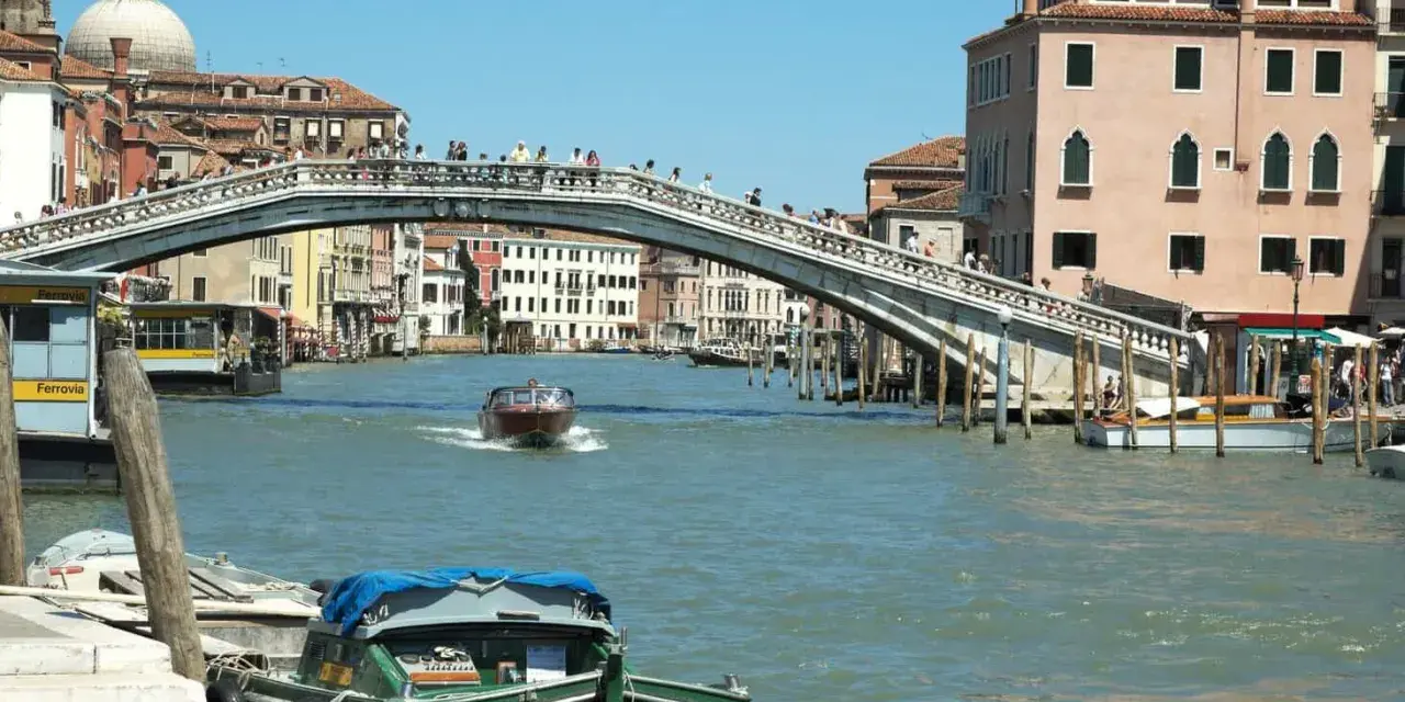 Un motoscafo naviga sotto uno dei ponti di Venezia, con edifici storici e gente che passeggia.