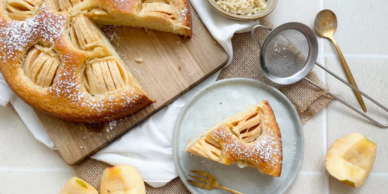 Ein köstlicher veganer Apfelkuchen, bestäubt mit Puderzucker, wird auf einem Holzbrett präsentiert. Daneben liegen frische Apfelstücke und eine Schale mit Mandelblättchen.