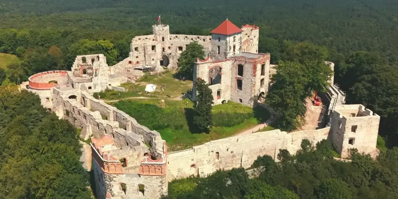 Ruiny zamku Tenczyn na wzgórzu, otoczone lasem. Widać mury obronne, wieże i pozostałości budynków.