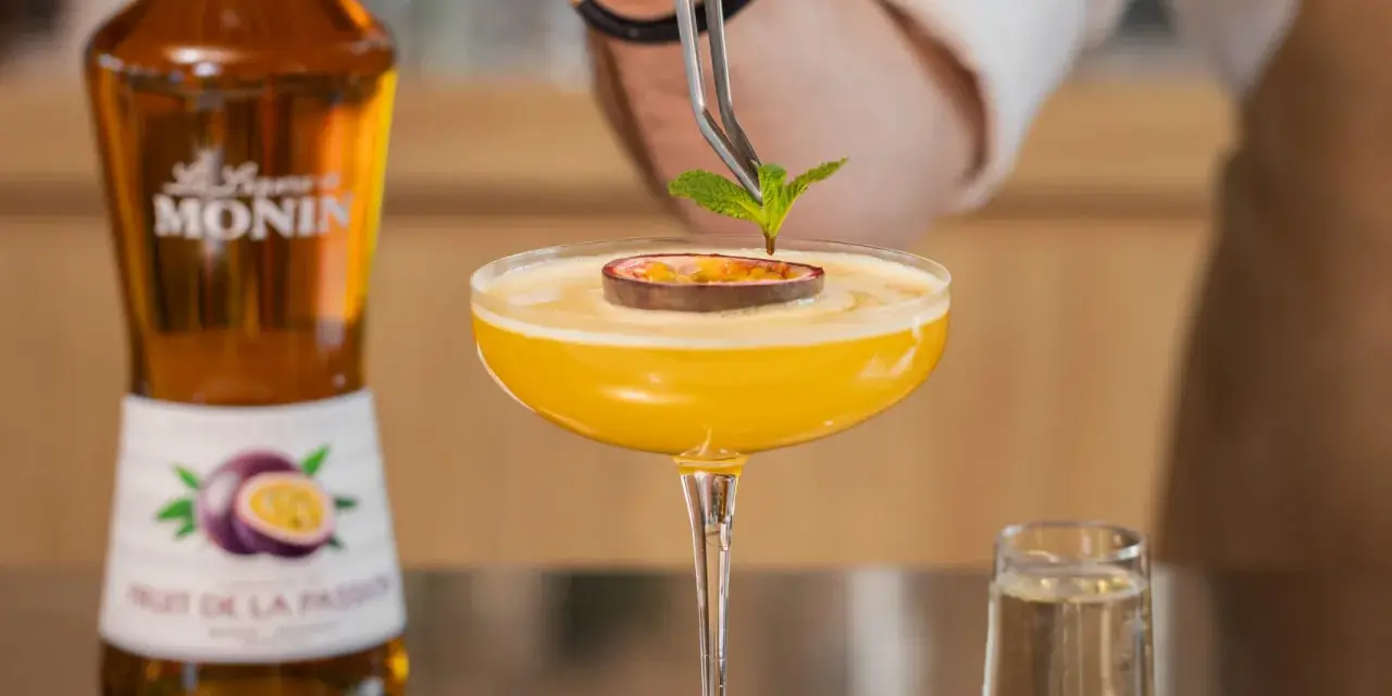 Barman dekoruje drink likierem z marakui, dodając listek mięty. Obok stoi butelka Monin.