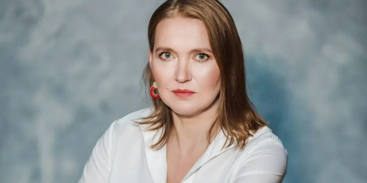 Kobieta w białej koszuli, z rudymi włosami i czerwoną biżuterią, udziela wywiadu. #16 Jak, krok po kroku, zrobić wywiad?
