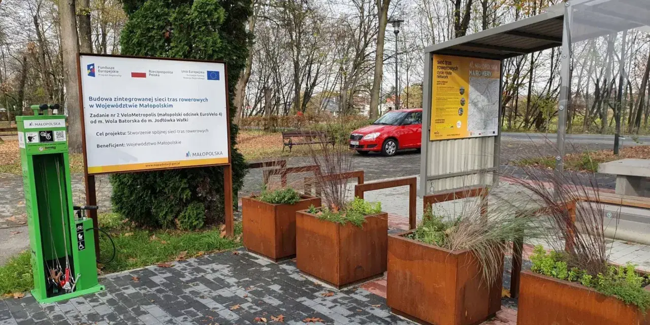 Zielona stacja serwisowa dla rowerów i tablica informacyjna o projekcie VeloMetropolis w Małopolsce. W tle czerwony samochód i jesienne drzewa.
