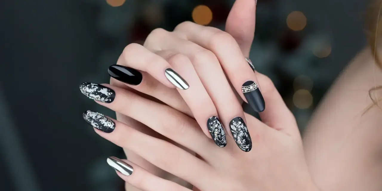 Mani con unghie particolari: nero lucido, argento specchiato e decorazioni floreali in bianco e argento con strass.