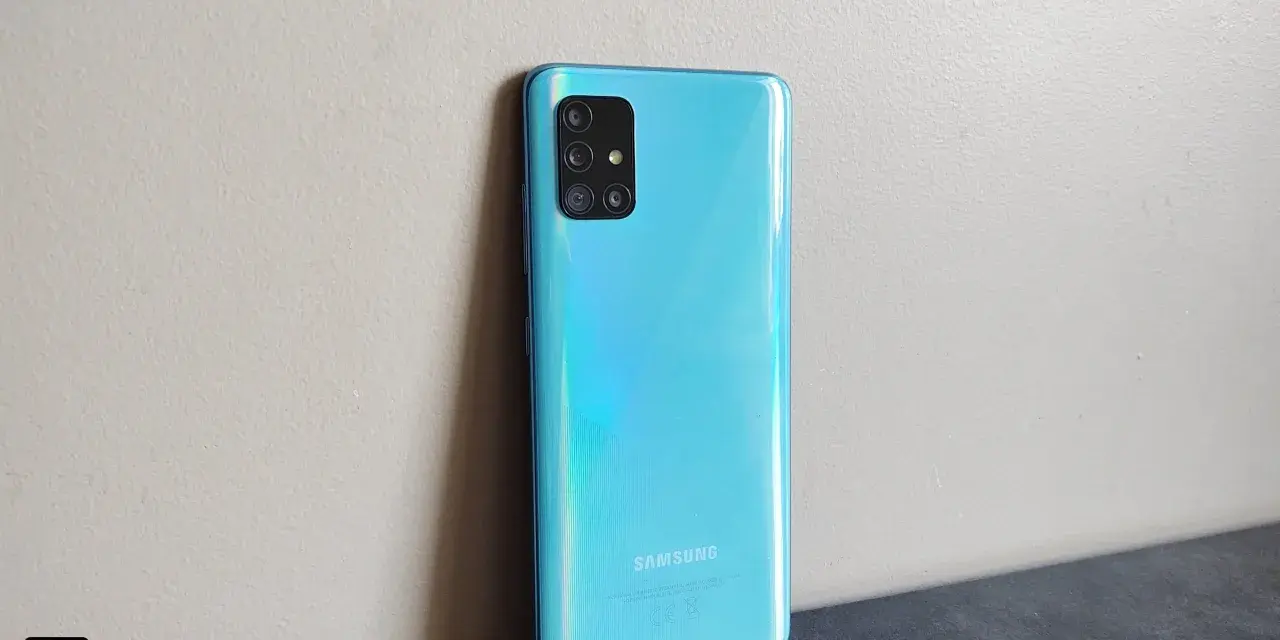 Błękitny Samsung A51 z aparatem w kwadratowej ramce, oparty o jasną ścianę.